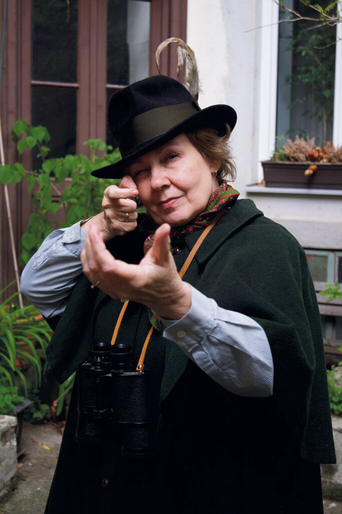 Straße der Frauen: Eine ältere Frau mit Hut und Fernglas um den Hals formt mit den Händen eine Geste, als würde sie zielen, im Innenhof vor einem Gebäude.
