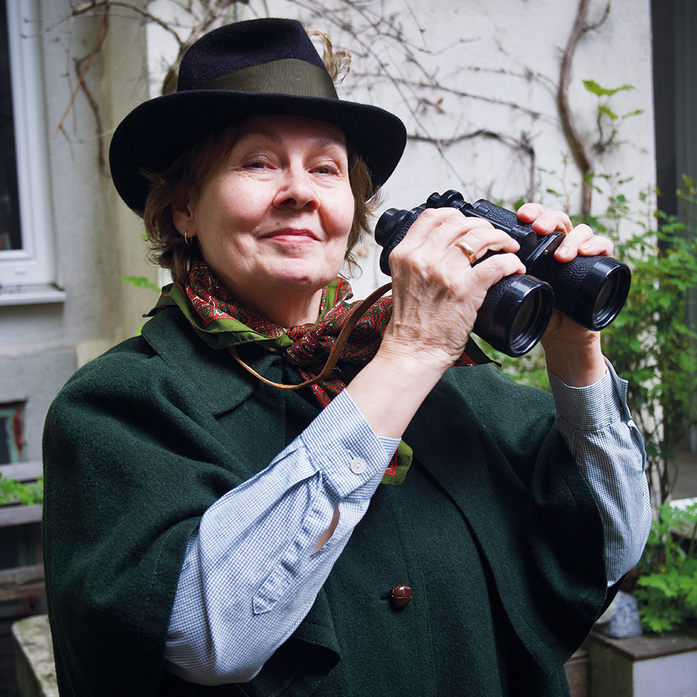 Straße der Frauen: Eine ältere Frau mit Hut hält ein Fernglas in den Händen und lächelt leicht, im Innenhof vor Fenstern und Kletterpflanzen.