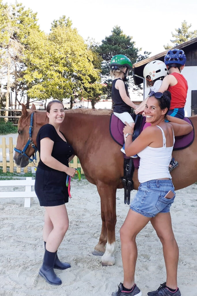 Zwei Frauen begleiten Kinder mit Helmen beim Reiten auf einem Pferd und lächeln in die Kamera auf einem Reitplatz.