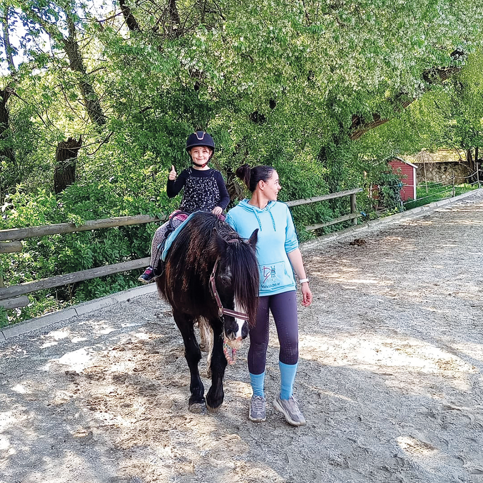 Llino: Ein Kind mit Reithelm sitzt auf einem Pony und zeigt den Daumen nach oben, während eine Frau das Pferd auf einem Reitplatz führt.