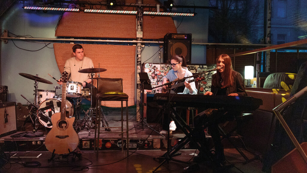 Sofia Reyna: Band spielt live auf kleiner Bühne mit Schlagzeug, Keyboard und Gesang in stimmungsvoll beleuchteter