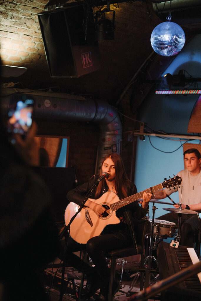 Sofia Reyna: Sängerin spielt Akustikgitarre und singt live auf einer Bühne, begleitet von einem Schlagzeuger im Hintergrund.