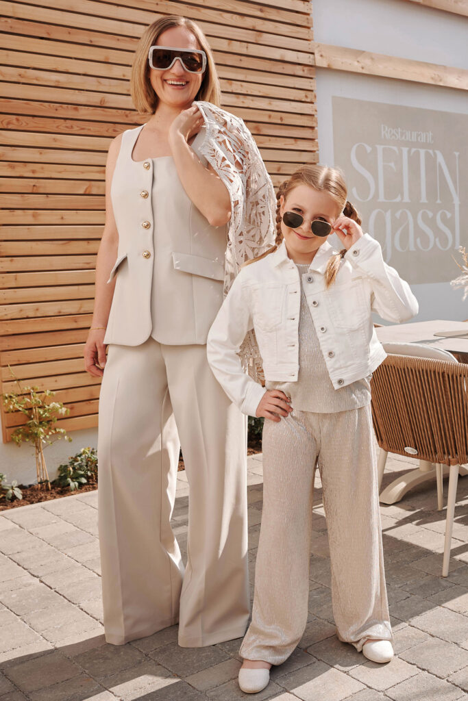Mutter-Tochter-Shooting: Mutter und Tochter posieren draußen vor einem Restaurant in hellen, abgestimmten Outfits und tragen Sonnenbrillen.