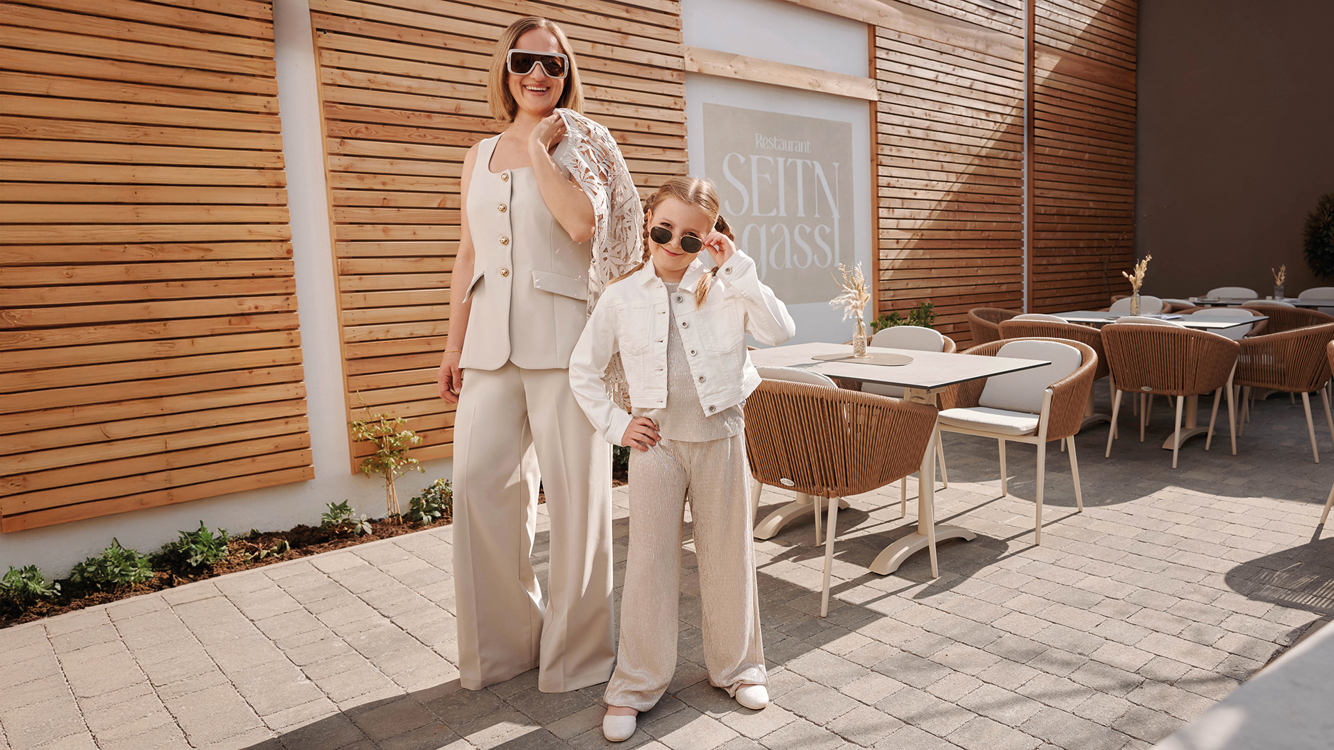 Mutter-Tochter-Shooting: Mutter und Tochter posieren draußen vor einem Restaurant in hellen, abgestimmten Outfits und tragen Sonnenbrillen.