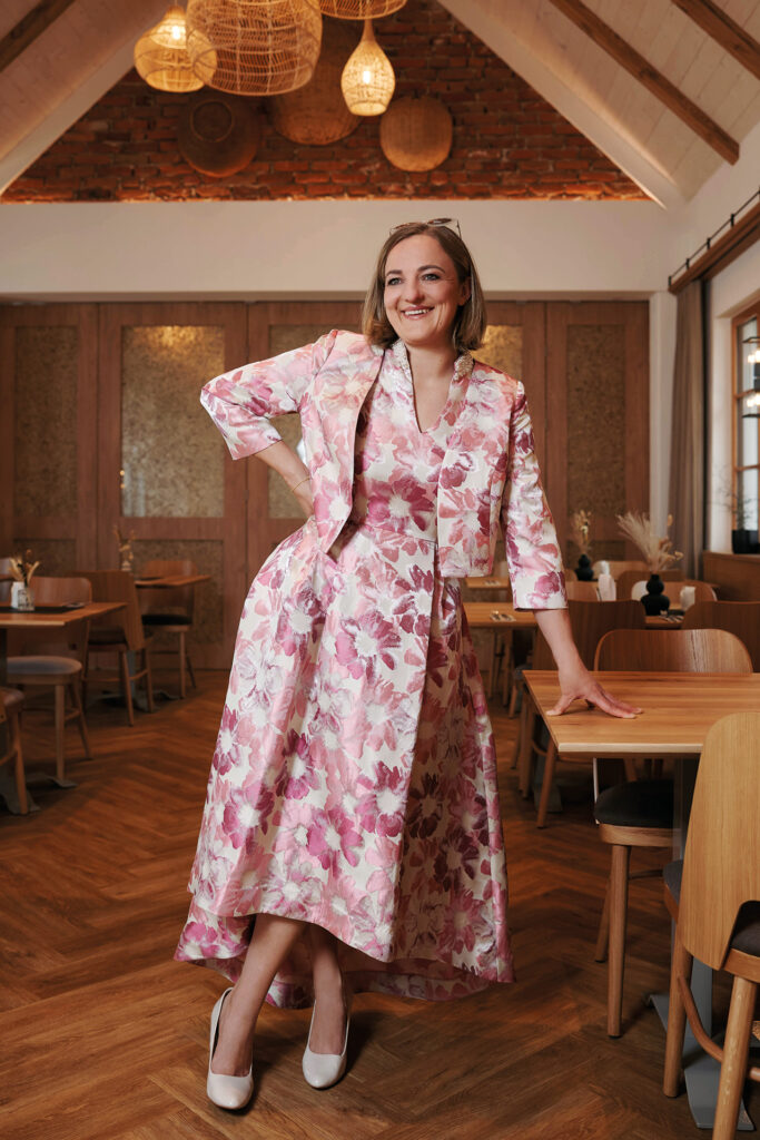 Mutter-Tochter-Shooting: Eine Frau in floralem Kleid steht in einem Restaurant und lehnt sich lässig an einen Tisch.