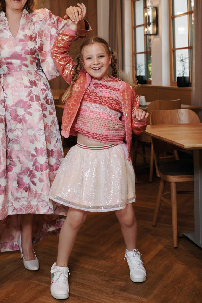 Ein Mädchen in rosa Outfit tanzt fröhlich und hält die Hand ihrer Mutter in einem Restaurant.