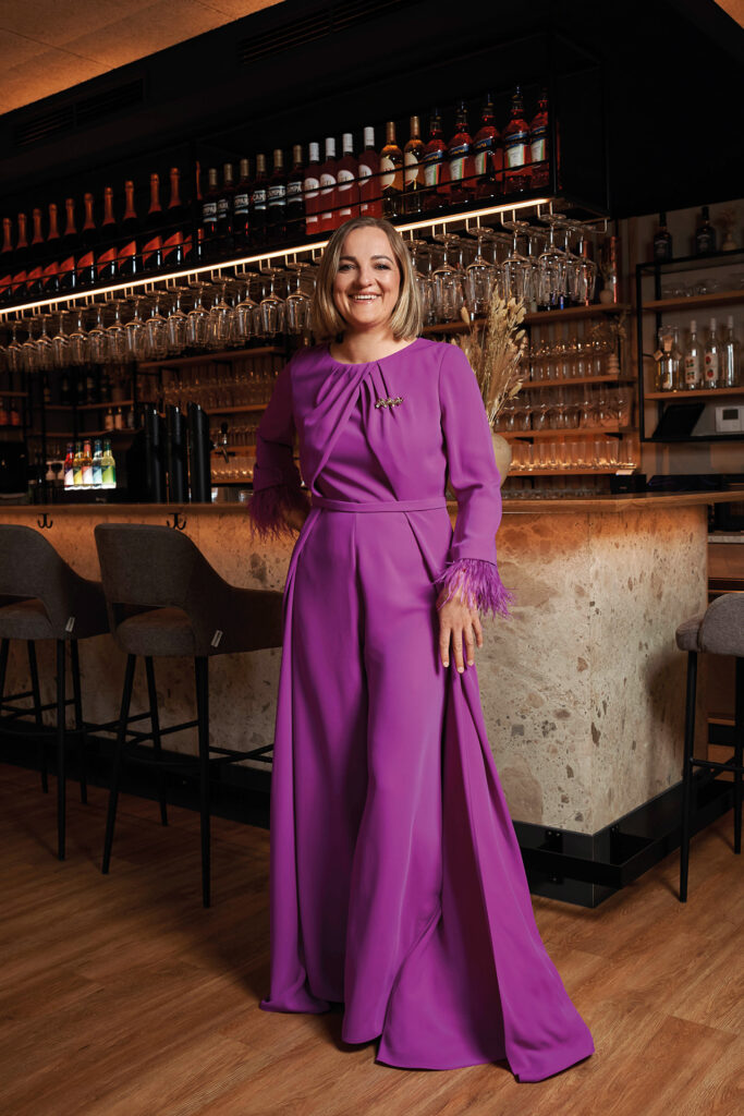 Mutter-Tochter-Shooting: Eine Frau in einem violetten Kleid steht an einer Bar und lächelt in die Kamera.
