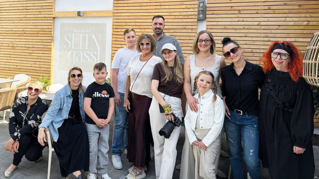 Eine größere Gruppe steht vor einem Restaurant, darunter auch eine Mutter mit ihrer Tochter, die gemeinsam in die Kamera lächeln.