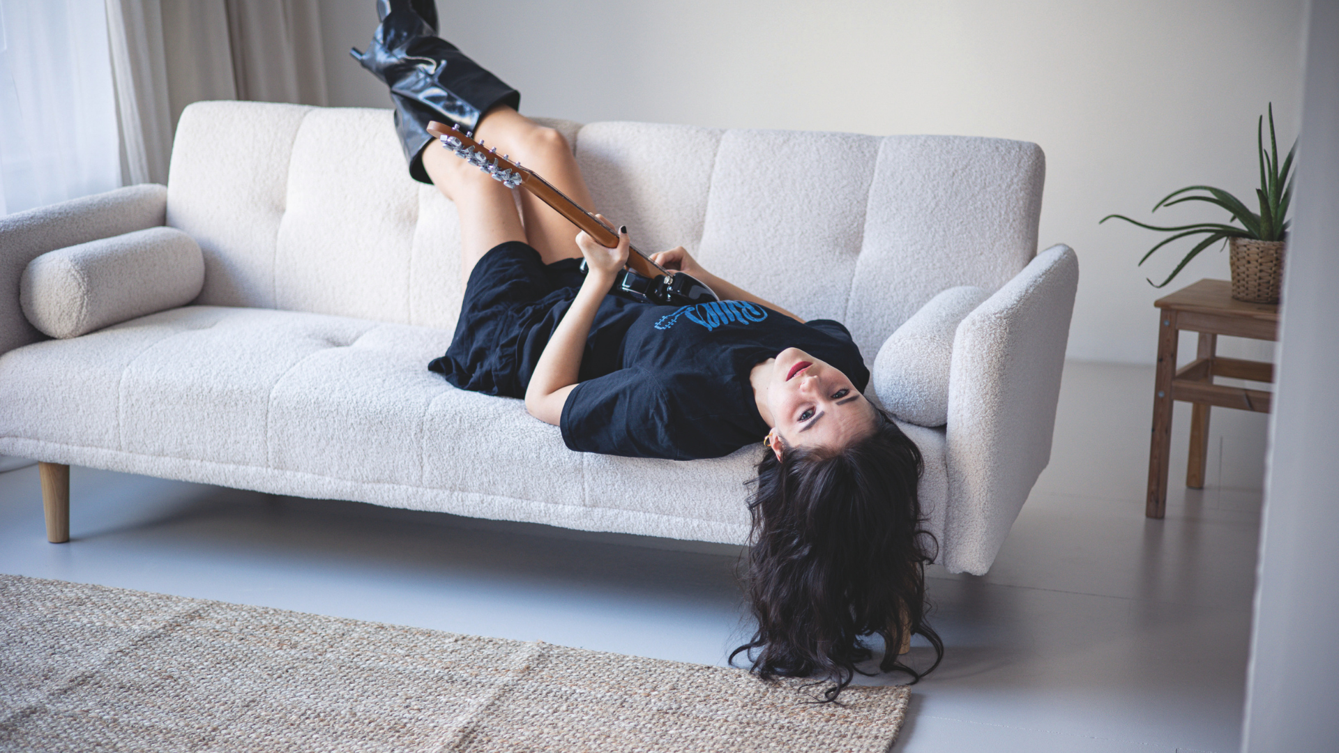 Lena Schaur liegt mit dem Rücken auf einem Sofa, mit einer Gitarre in der Hand und dem Kopf nach unten hängend und sieht direkt in die Kamera