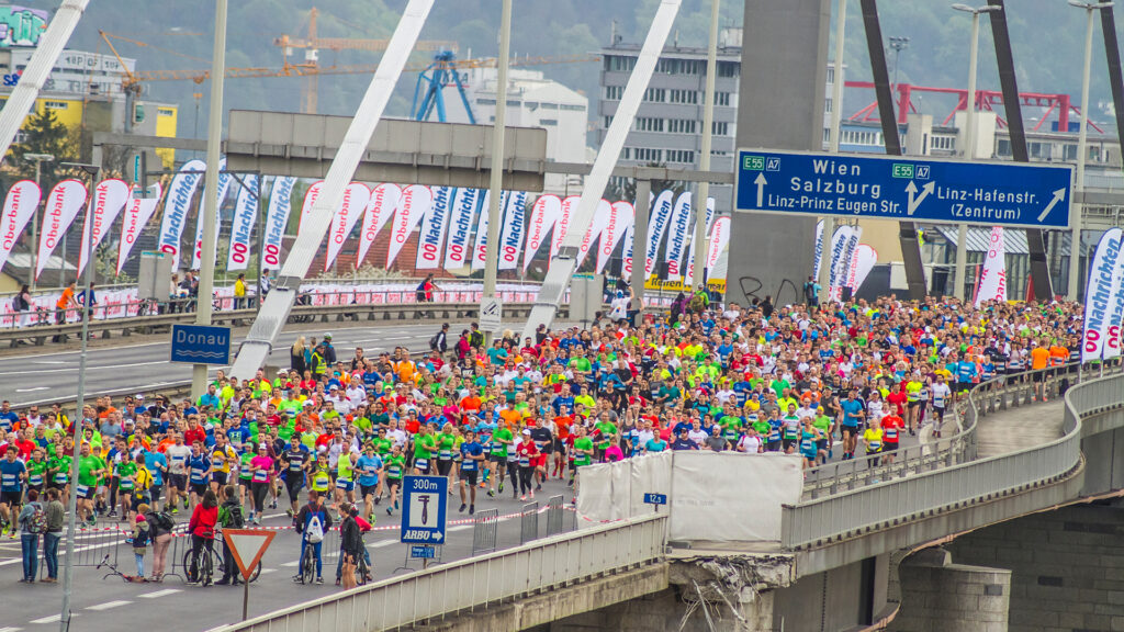 Events Österreich: Große Teilnehmergruppe beim Linz Marathon auf einer Brücke mit Blick auf Stadt und Beschilderung.