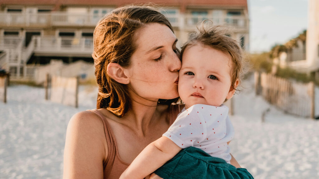 Mutter küsst ihr Kleinkind am Strand bei warmem Abendlicht, während das Kind in die Kamera blickt.