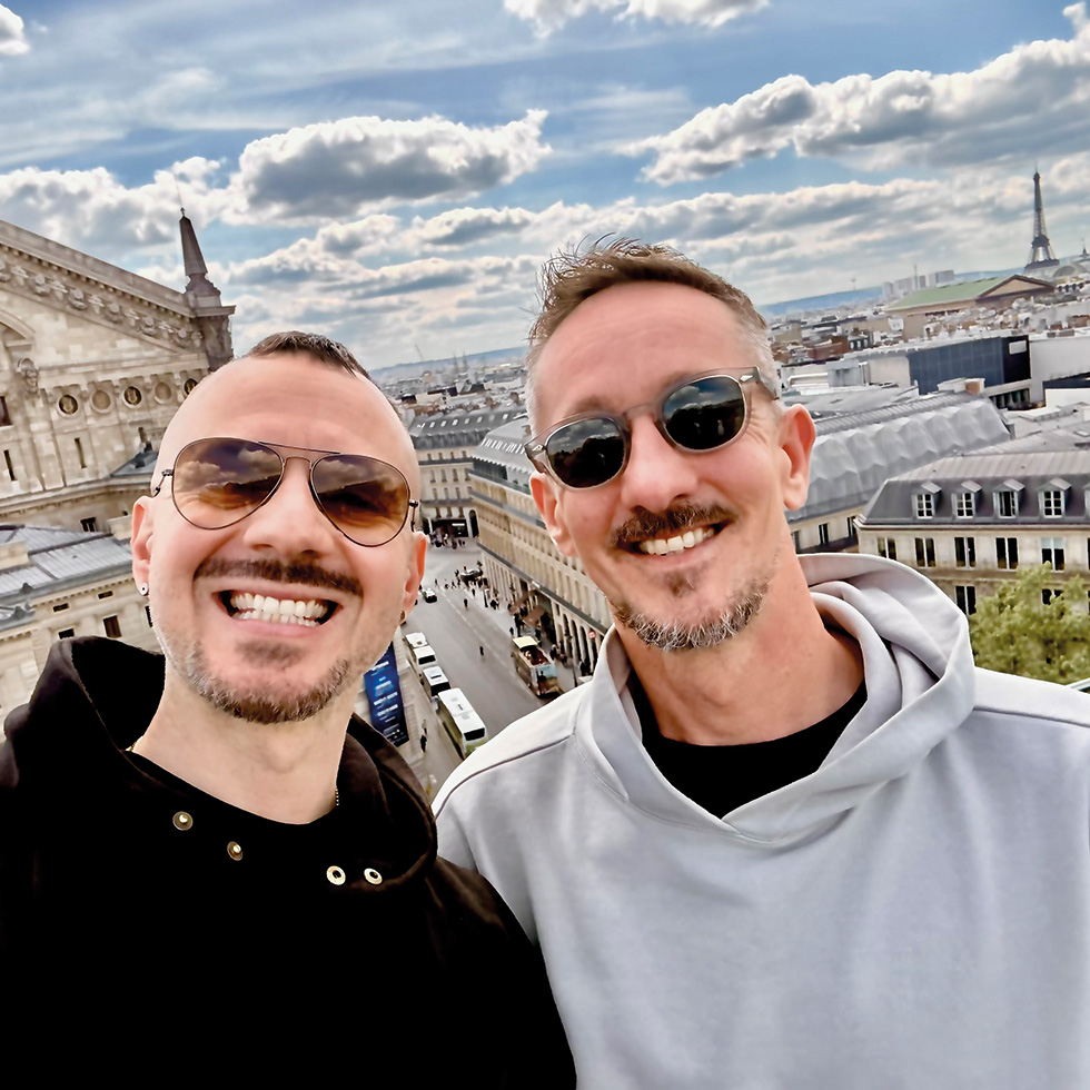 Zwei lächelnde Männer mit Sonnenbrillen vor der Skyline von Paris auf einer Dachterrasse.