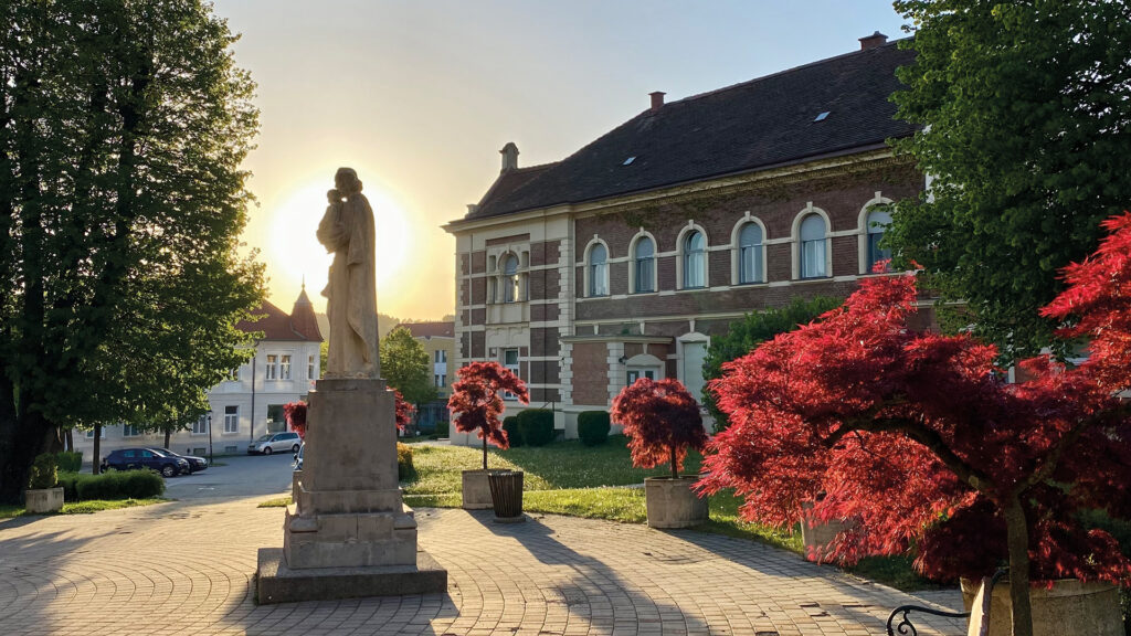 Pinkafeld im Abendlicht: Josefipark mit Statue, historischem Gebäude und rot leuchtenden Bäumen bei Sonnenuntergang.