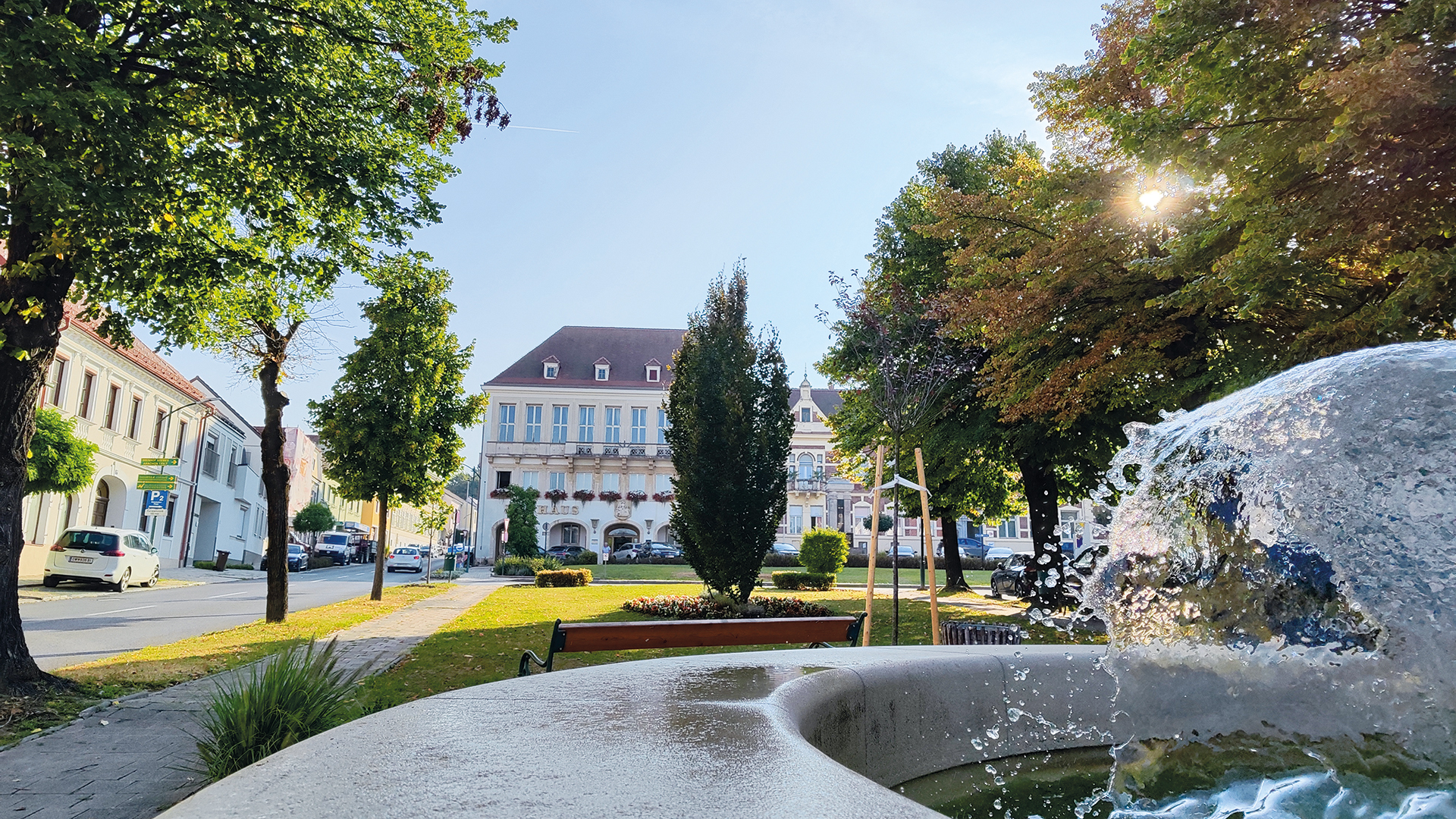 Pinkafeld Stadtzentrum: Brunnen, Rathaus und grüne Parkanlage im sommerlichen Morgenlicht.