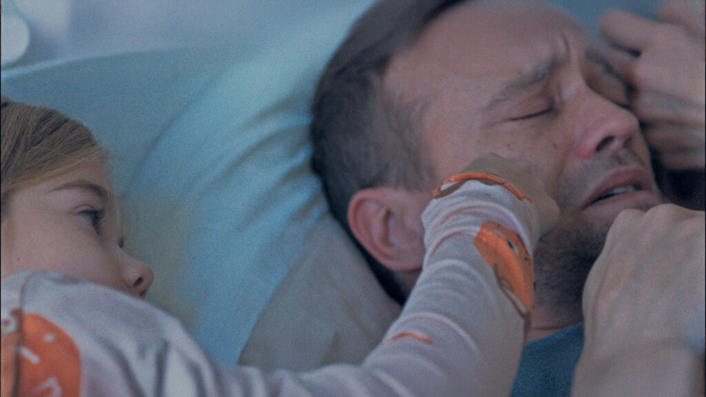 Vater liegt erschöpft im Bett, während seine Tochter ihn aufmerksam ansieht – Szene aus dem Kurzfilm „Bester Papa, manchmal“.