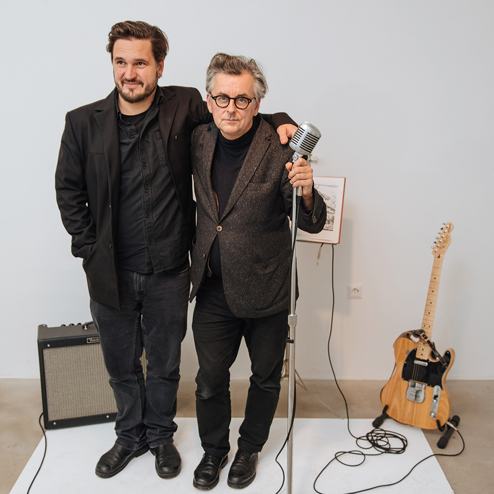 Events Juni: Zwei Männer stehen gemeinsam vor einem Mikrofon, daneben Verstärker und E-Gitarre in einer reduzierten Studio-Szene.