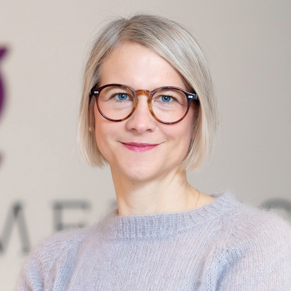 Hack your Health: Porträt einer blondhaarigen Frau mit Brille und hellem Strickpullover vor neutralem Hintergrund.