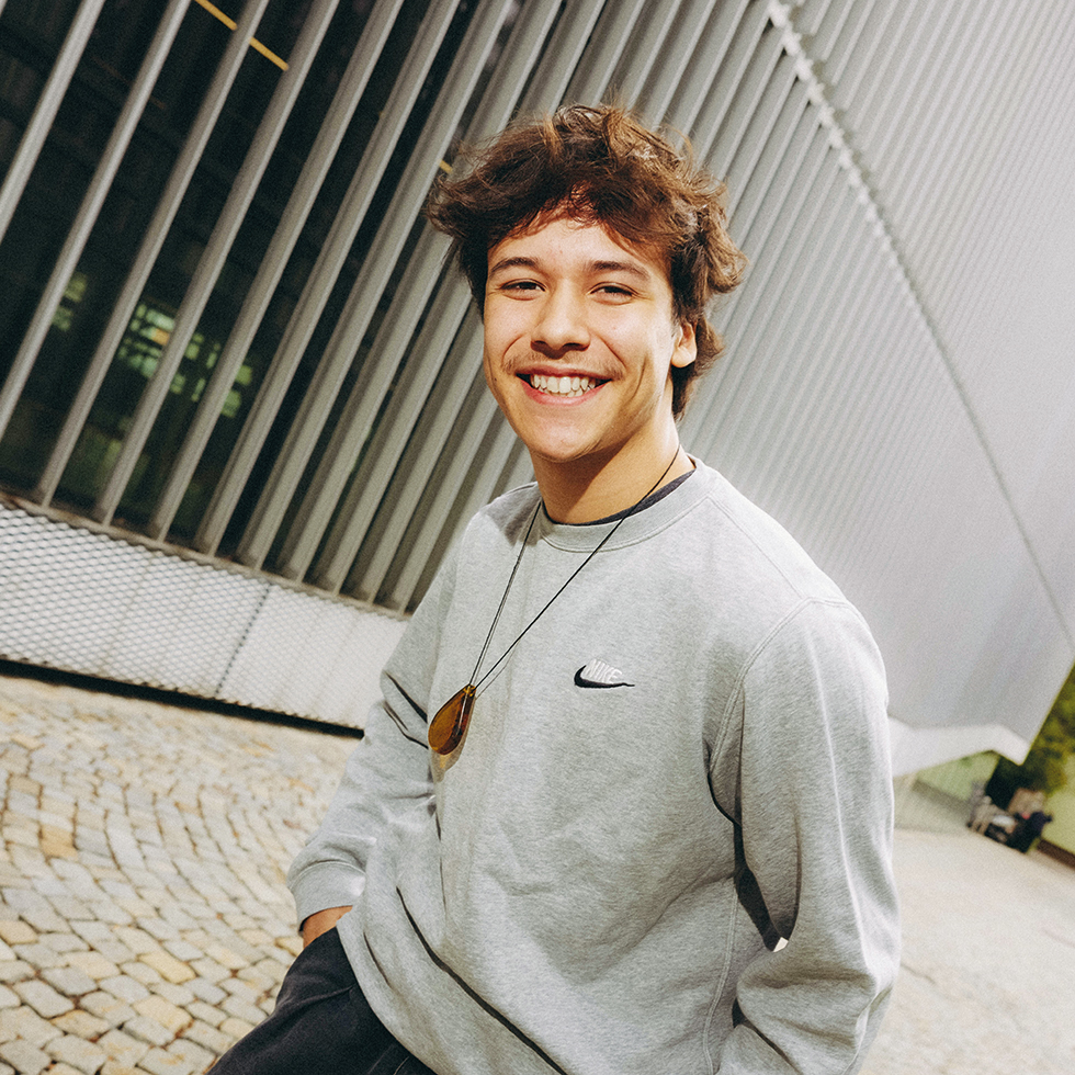 Churro64 lächelt bei einem Streetstyle-Porträt vor moderner Architektur in die Kamera.
