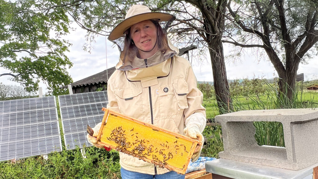 Julia Mainx in Imkerschutzkleidung mit einer Honigwabe bei der Arbeit an ihren Bienenstöcken im Grünen.
