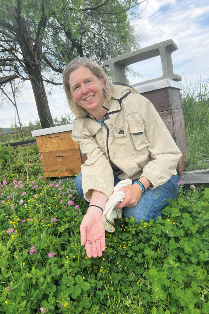 Julia Mainx sitzt neben ihren Bienenstöcken auf einer Blumenwiese und hält eine Biene auf der Hand.