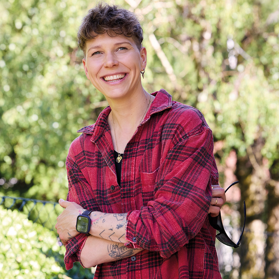 Virginia Ernst bei einem entspannten Outdoor-Porträt mit kariertem Hemd und sommerlicher Naturkulisse.