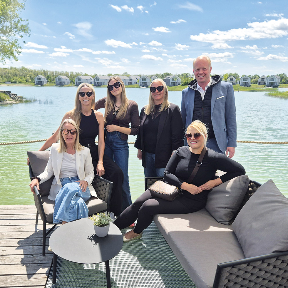 Sommer-Teamfoto am See: Gruppe sitzt und steht auf einer Terrasse direkt am Wasser vor modernen Ferienhäusern.