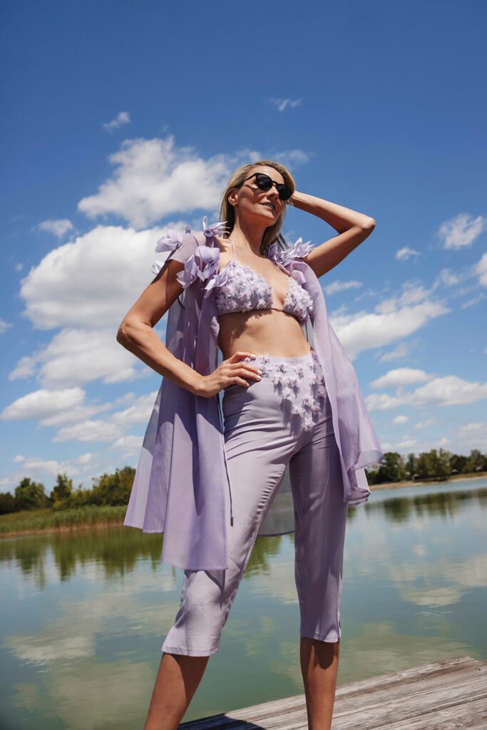 Sommer-Couture am Wasser: Frau in fliederfarbenem Fashion-Outfit mit floralem Design auf einem Steg am See.
