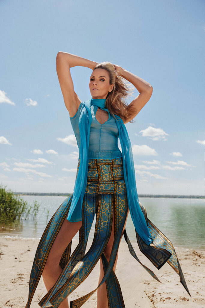 Sommer-Fashion am Strand: Frau in türkisem Outfit mit wehenden Stoffbahnen am Seeufer.