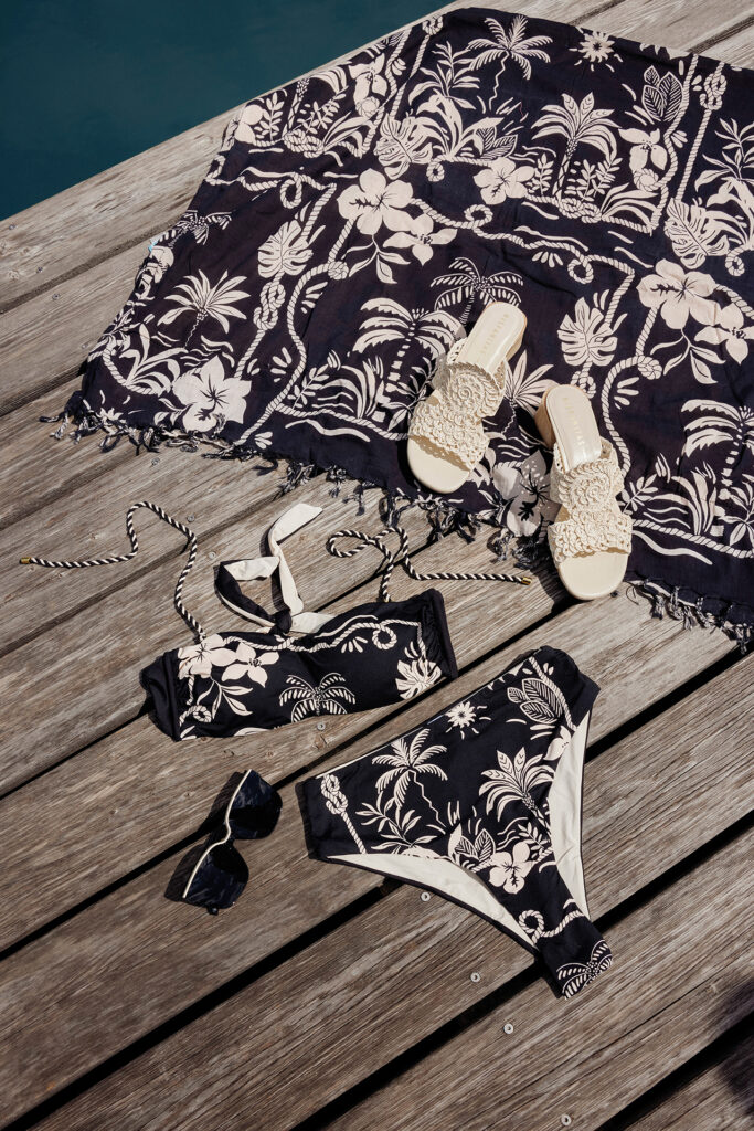 Sommer-Bikini mit tropischem Muster, Sonnenbrille und Sandalen dekorativ auf einem Holzsteg arrangiert.
