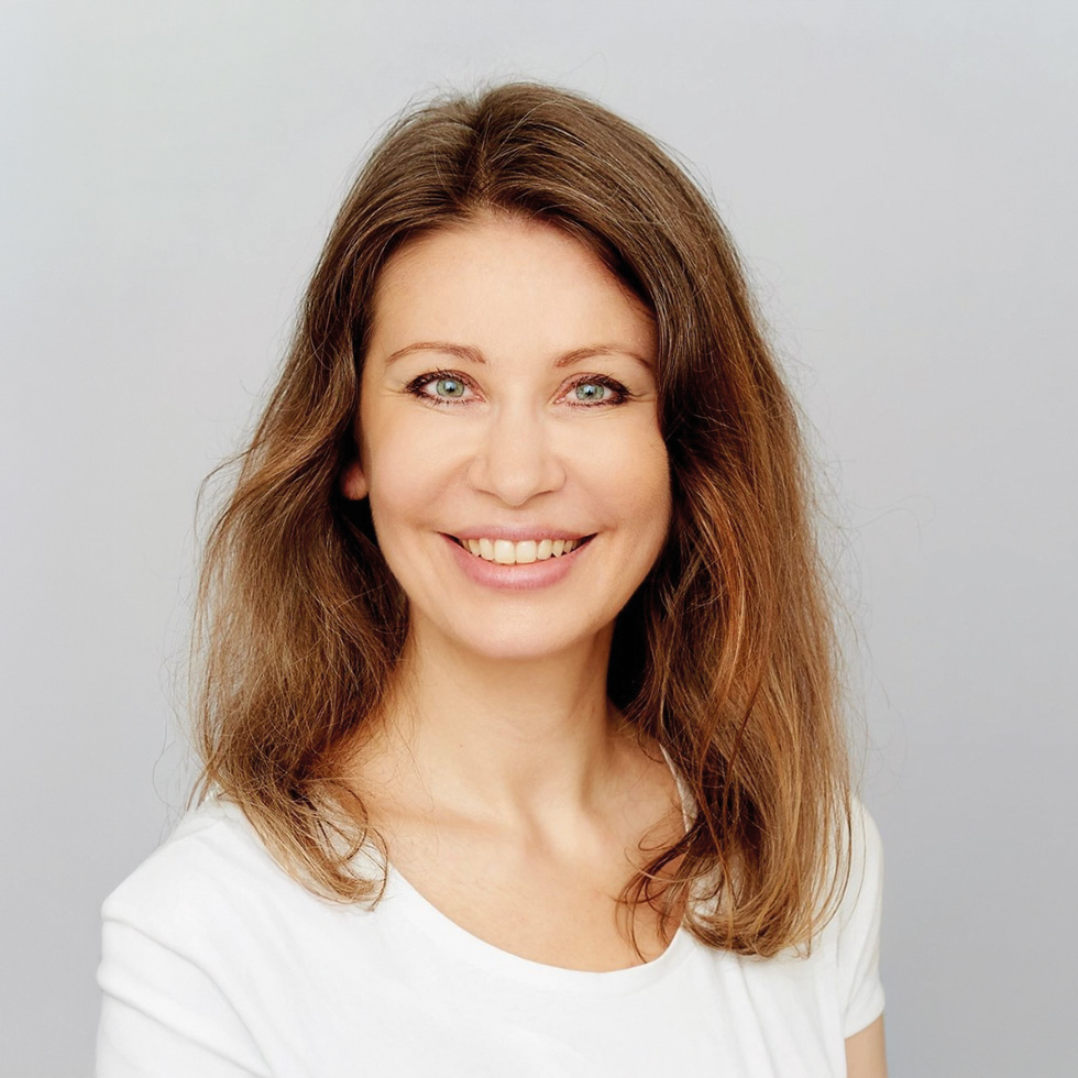 Hack your Health: Porträt einer lächelnden Frau mit braunem Haar und weißem Shirt vor hellem Hintergrund.