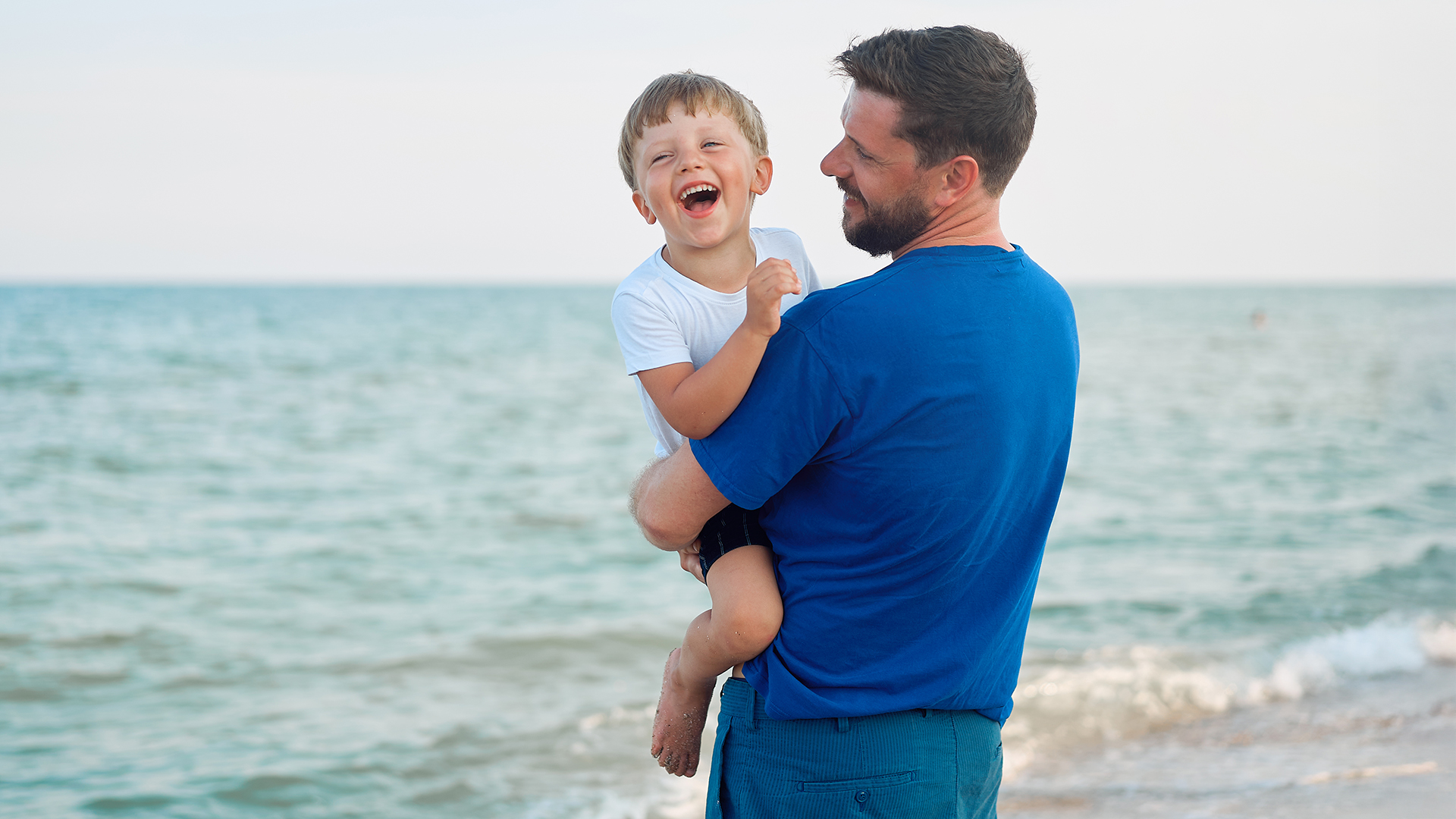 Vater trägt lachenden Sohn am Strand auf dem Arm und genießt gemeinsame Zeit zum Vatertag am Meer.