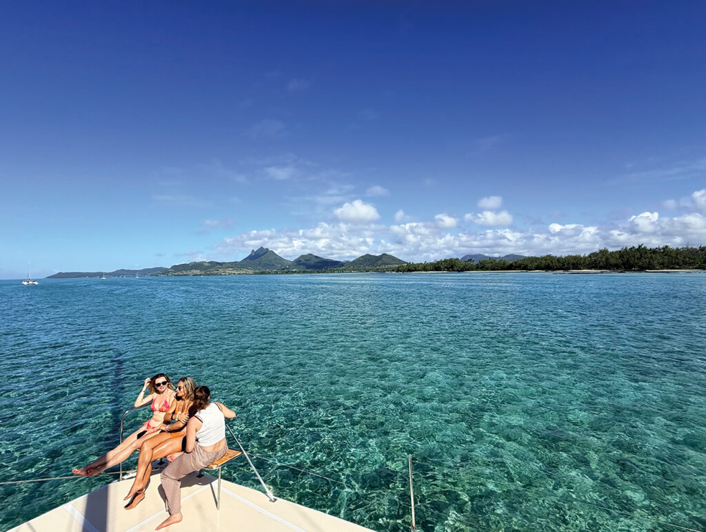 Mauritius: Drei Frauen genießen auf einem Katamaran die Aussicht auf das türkisfarbene Meer und die Küste von Mauritius.