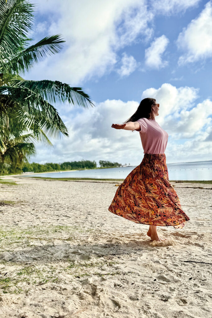Mauritius: Frau dreht sich barfuß am weißen Sandstrand vor Palmen und Lagune auf Mauritius.