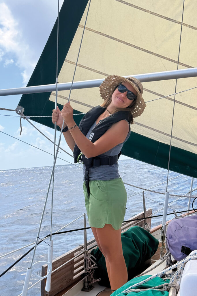 Absolventin auf dem Atlantik: junge Frau beim Segeln mit Rettungsweste und Sonnenhut.