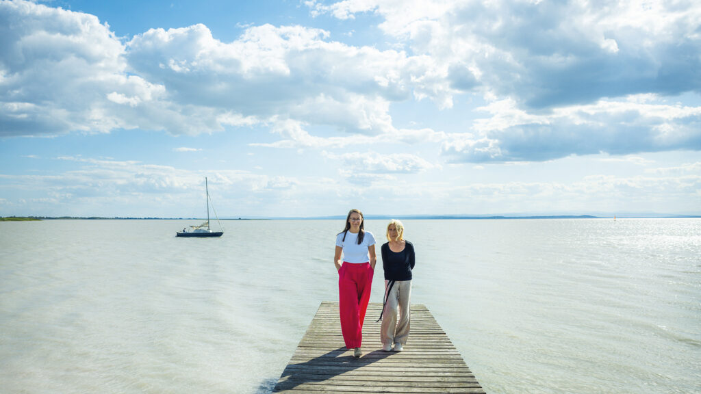 Nincshof am Neusiedler See: Zwei Frauen auf einem Holzsteg vor weiter Seelandschaft und Segelboot.