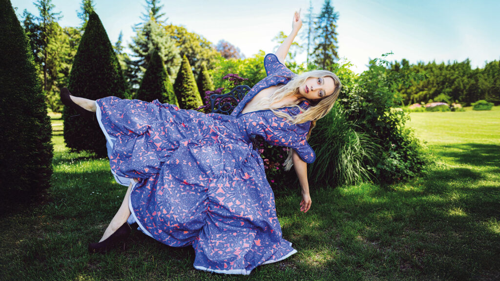 Selina Heindl bei einem Designershooting in einem weit geschnittenen gemusterten Kleid in einem grünen Garten.