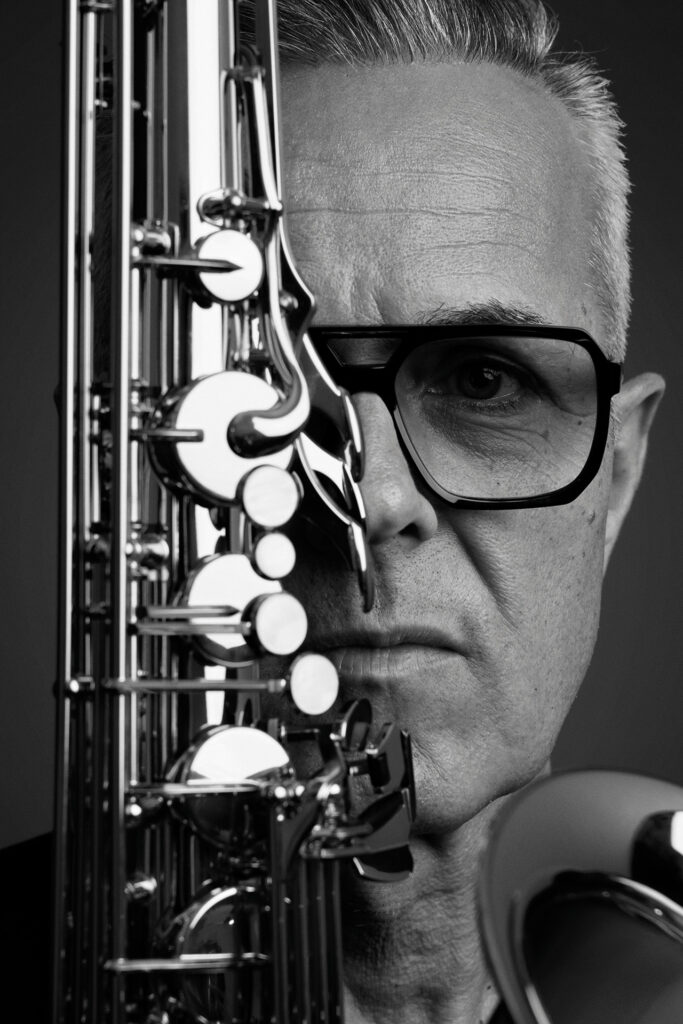 tomX im Schwarz-Weiß-Porträt mit Saxofon und markanter Brille.