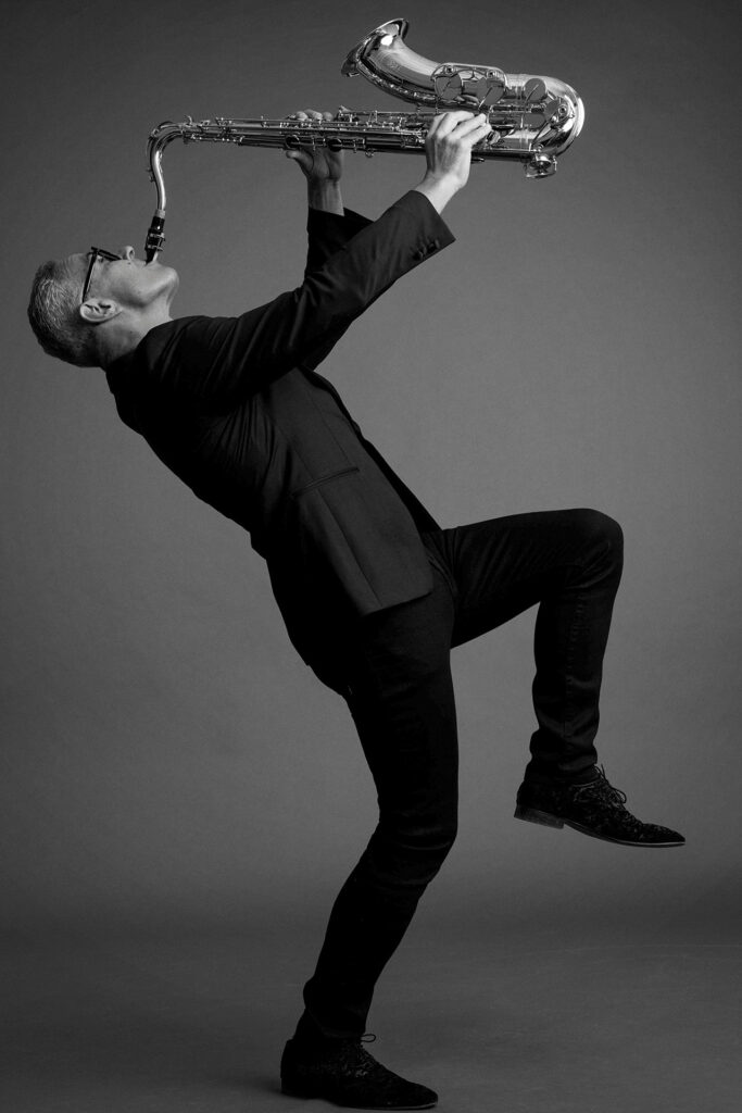 Saxofonist tomX spielt sein Instrument in dynamischer Pose im Schwarz-Weiß-Studio.