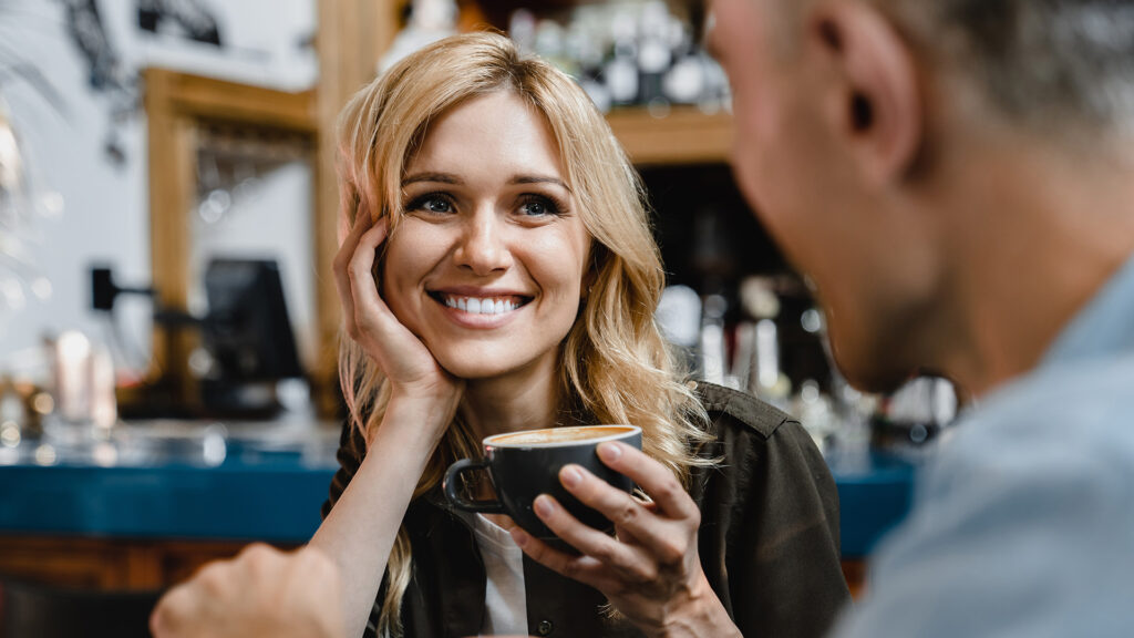 Flirten: Lächelnde Frau flirtet bei einer Tasse Kaffee mit einem Mann und sucht seinen Blick.